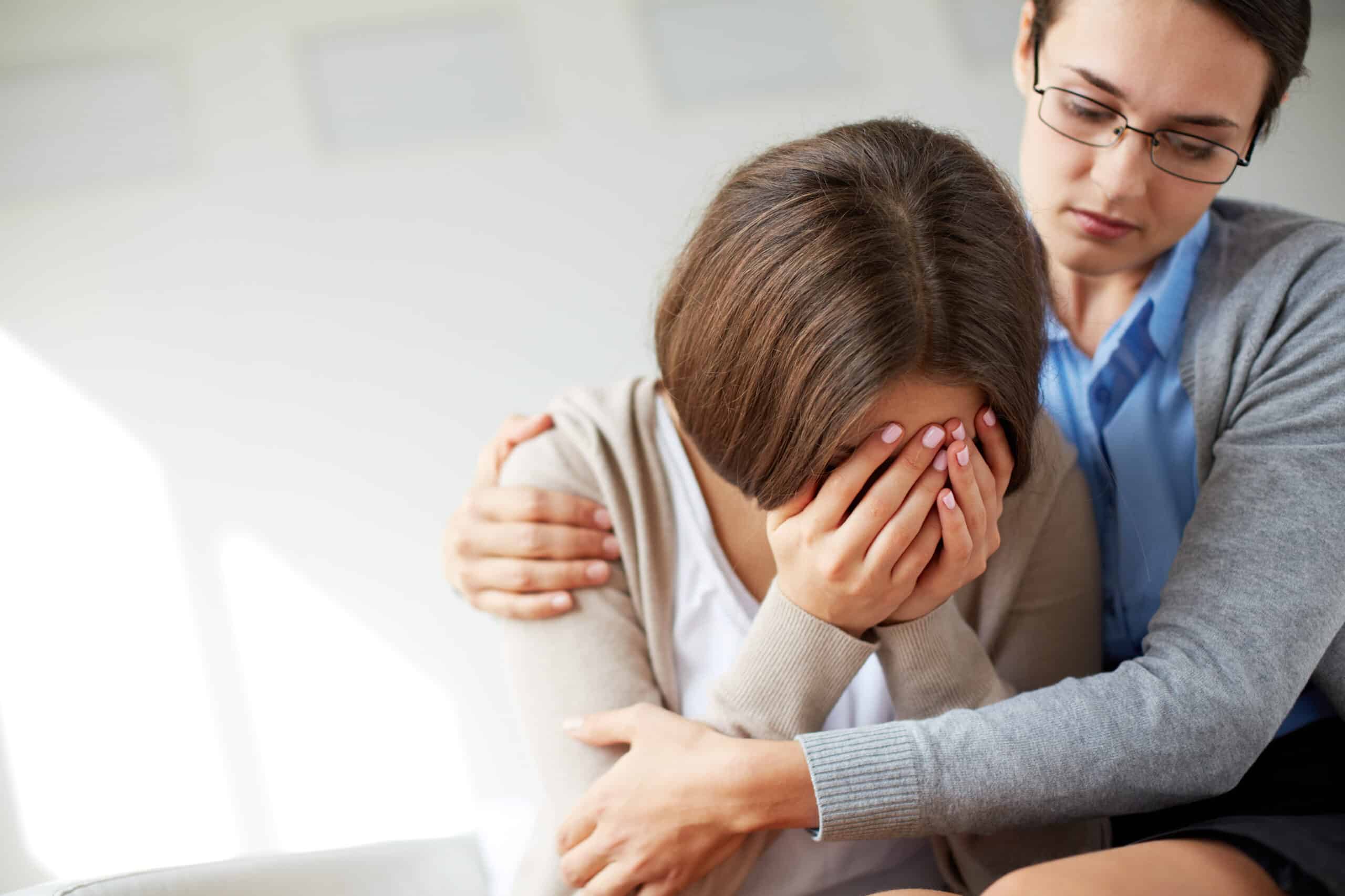 Picture shows two women. The young woman on the left is despaired covering her face with hands. The woman on the right hugs her. The background of the picture is grey. The picture refers to grief counselling.