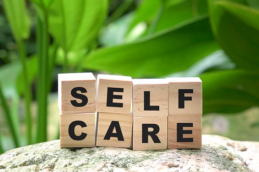 Self-Care for physical and mental health. The words self and care written on wooden blocks on top of each other.  The setting is a rock with diffuse green leaves in the background.  The words self care for physical and mental health are written below the picture.