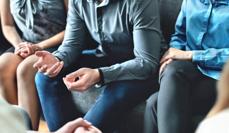 A group of 5 men and women closely and comfortably sitting together.  The faces and feet are not in the picture and they are seated on a grey sofa.  This image expresses the mutual support that is a necessary part of relapse prevention.  