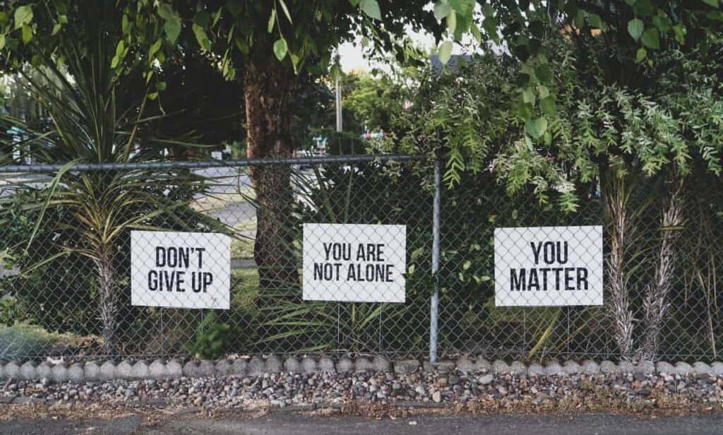 A chain linked fence against the background of trees.  The fence shows 3 signs.  From left to right, One:  Don't give up, Two:  You are are not alone, and Three:  You matter.  These words of encouragement show that relapse prevention is possible, that there is support, and that the addict is a valuable person.  