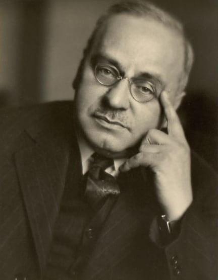 An old fashioned black on white picture of Dr. Alfred Adler.  In the picture he is wearing round metal glasses and is sporting a mustache.  His head is tilted to the side and he is pointing his index finger to his temple.  He is wearing a dark suit, vest, and tie over a white shirt.  This image reflects Adler's biography.