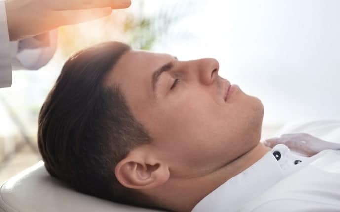 A young man with brown short hair wearing a white shirt prostrate on a treatment table.  His eyes are closed, and a hand is over his head.  The person who is holding the hand over the patient's head is in a white lab coat.  Bright light is showing between the hand and the young man's head.  This is showing hypnosis for smoking cessation