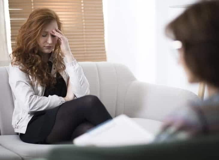 Treatment with Cognitive Behavioral Therapy (CBT) for panic attacks. A woman sitting on the sofa, holding her hand on the left site of the face. The psychotherapist is sitting on the right site writing notes. 