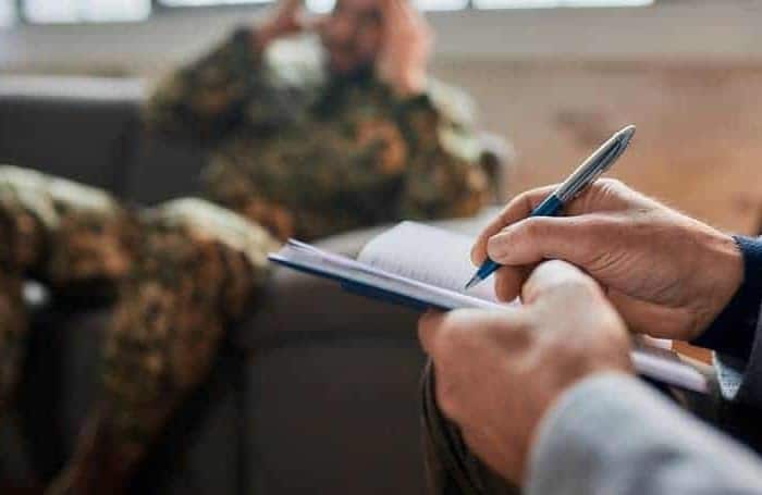 Psychotherapy (counselling) for PTSD in Dubai. The picture shows the psychotherapy treatment for PTSD. The soldier in a field uniform is half lying on the sofa. In front of him we see the arms of the psychotherapist holding a pen and making notes
