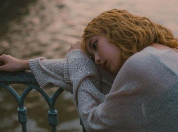 depression diagnosis treatment Dubai. The picture shows a young woman with her right arm on the blue iron railing of a bridge. In the background we a river. The woman is wearing a light blue pullover. Her face expresses sadness and worries. The photograph captures the main signs of depression such as sadness, sorrow, low energy, resignation, and low self-esteem.