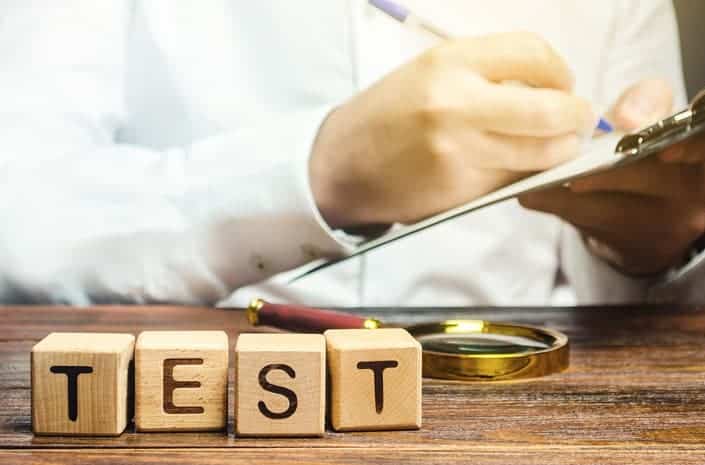 The picture shows a person holding a note and a pen, sitting behind a wooden desk. On the desk we see a magnifying glas and wooden cubes with the inscription: "test". The picture refers to psychometric testing